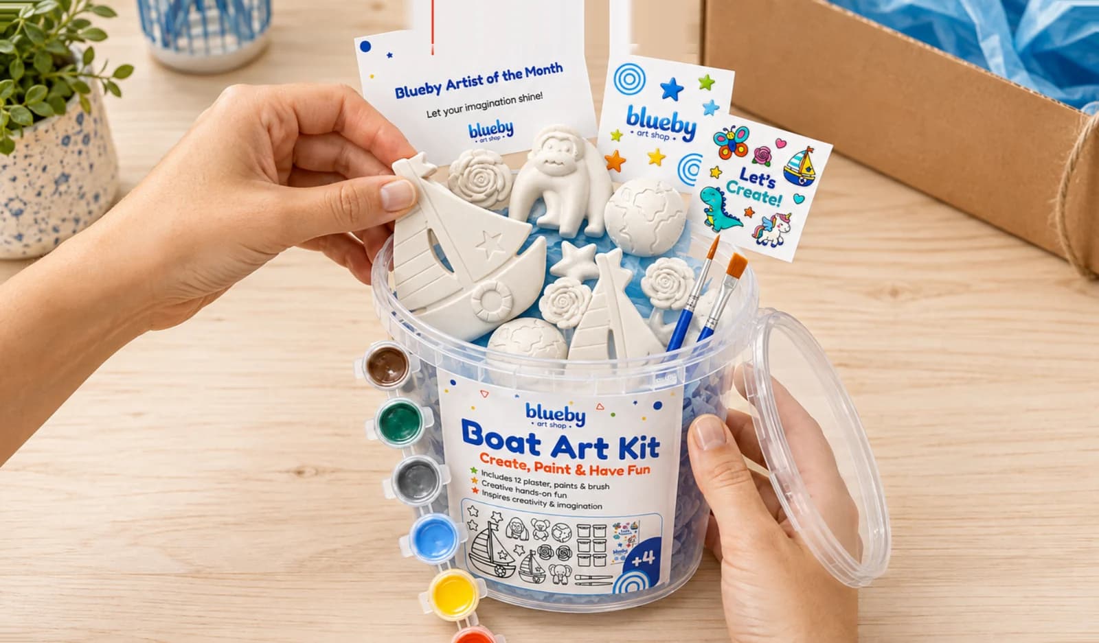 Hands packing a Blueby Boat Art Kit bucket with plaster pieces, paints and a brush